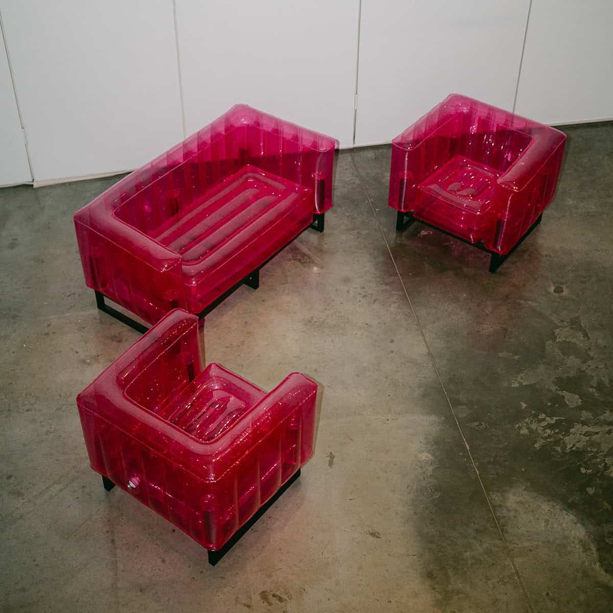 Three transparent hot pink inflatable lounge chairs arranged on a concrete floor against a white wall.
